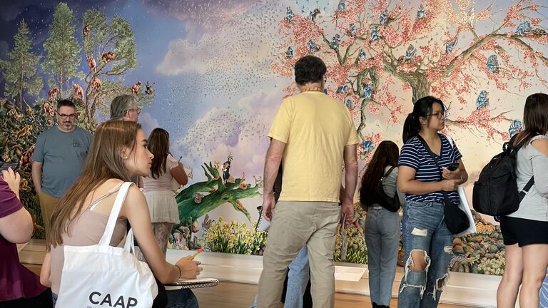 People of various ages, ethnicities, and genders viewing a mural. A person in the foreground has a tote that reads "CAAP. College Arts Access Program."