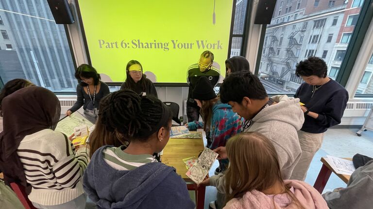 Students of varying genders and ethniticies gather around a table. A projection on a screen in the background reads: "Part 6: Sharing Your Work."