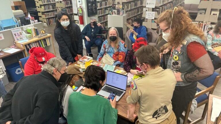 People of different ages sit and stand around a table in a public library, looking at a laptop and phones during an intergenerational LGBTQ+ gathering.
