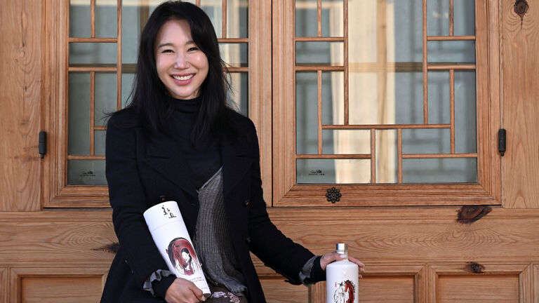 A Korean woman with long styled brown hair sits in front of a light blond wood door with traditional Korean firmaments in the window. She holds Hwayo soju bottles in her hand (resting on the out of view bench she sits on) and in her lap. 