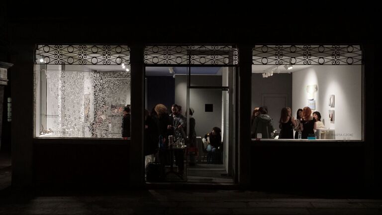 The illuminated storefront of the Czok Foundation at night, viewed from the street. Through large windows and an open door, visitors mingle inside the gallery, with Anya Lomachenko's intricate textile work Undersky visible in the left window.