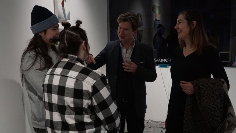 Four people in conversation at the Undercurrents opening reception inside the Czok Foundation gallery. Two students laugh with a man in a blazer and a woman smiling beside him, with artwork visible on the walls behind them.
