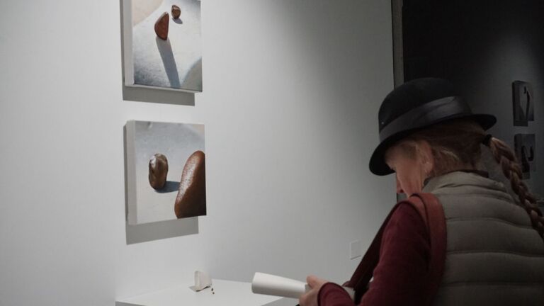 A visitor wearing a black bowler hat and red jacket reads an exhibition pamphlet while viewing two small square paintings mounted vertically on a white gallery wall. The paintings depict smooth brown stones in stark light and shadow. A small sculptural object sits on a pedestal below them. The work is The Midway by Younsoo and Jack Flynn.