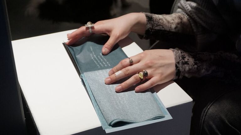 A close-up of two hands with rings turning the pages of a blue-gray artist's book resting on a white illuminated surface. The pages contain printed text and translucent paper layers.