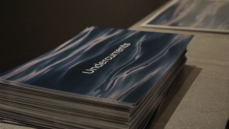 A stack of exhibition catalogs on a flat surface, their covers featuring a close-up photograph of deep blue-gray rippling water with the word "Undercurrents" set in white type. Additional printed materials are partially visible in the background.