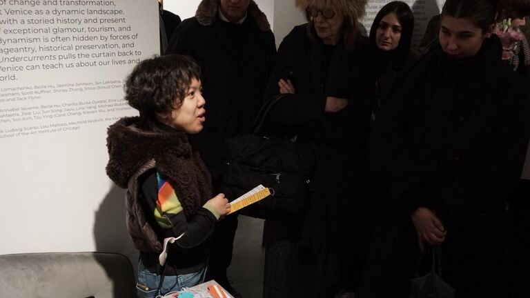 A student holds a small yellow card and speaks to a group of attendees gathered in front of the Undercurrents exhibition wall text, which lists participating artists and collaborators. Colorful tags from Shirley Zhong's Wheelchair Book are visible on a table in the foreground.