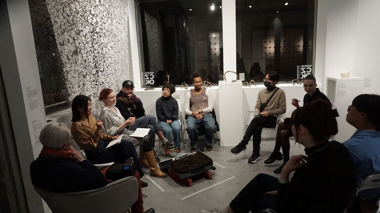 A group of approximately twelve people seated in a circle of chairs inside the Czok Foundation gallery, engaged in conversation during an artist talk. Anya Lomachenko's Undersky textile is visible on the wall behind them, alongside Rome Wareham's bronze sculptures on white pedestals. Jasmine Johnson's open suitcase installation sits on the floor at the center of the circle.