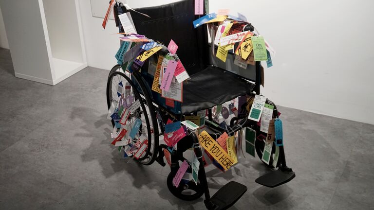 A black wheelchair covered in dozens of colorful paper tags and luggage labels of varying sizes, shapes, and colors. Visible text on the tags includes words like "Assistance," "Standby," "Late Check-In," "Are You Here," and "Danger." The work sits on a concrete floor against a white gallery wall.