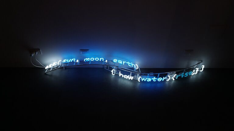 A neon light sculpture mounted low on the floor in a darkened room. The glowing blue and white text reads "(sun, moon, earth)" on one undulating line and "{ how (water)(rise) }" on a second line below, with the tubing forming a wave-like shape that casts a blue glow on the surrounding floor and wall.