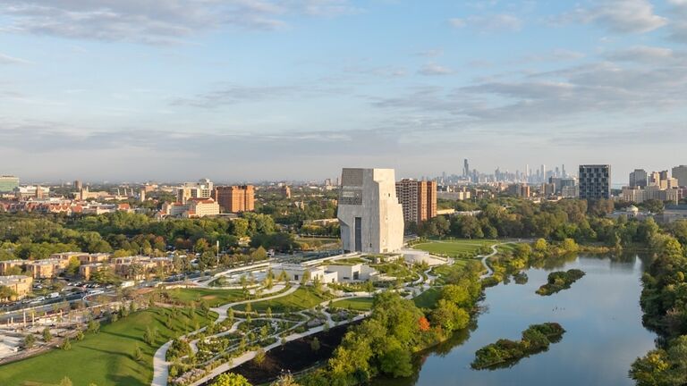 Aerial view of the Obama Presidential Center campus in Jackson Park, with the museum tower rising above surrounding greenery, a lagoon in the foreground, and the Chicago skyline visible in the distance.