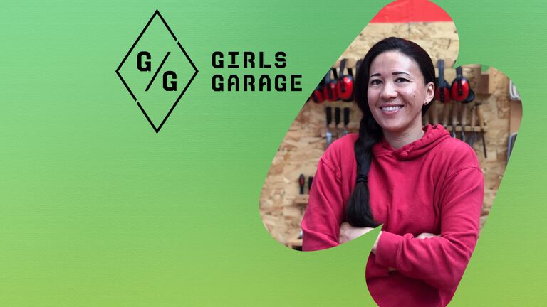 Emily Pilloton-Lam stands with arms crossed, smiling, in front of a wall of tools inside the Girls Garage workshop. The Girls Garage logo appears to the left against a green background.