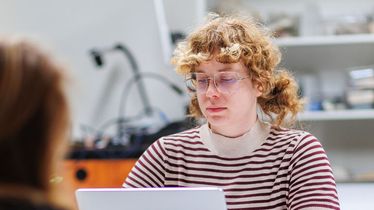 A person with light skin tone, glasses, and light curly hair at a desk with a laptop computer.