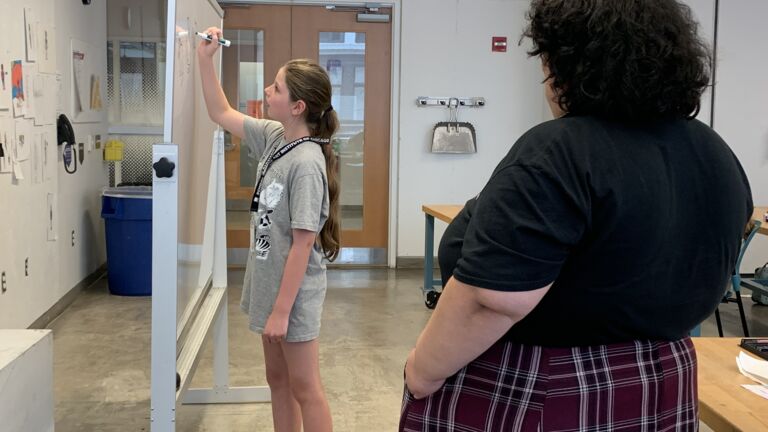 An instructor observes a student writing on a whiteboard. 
