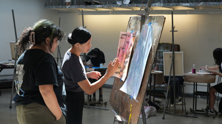 Two people with fair skintones looking at an easel and conversing in a classroom. 