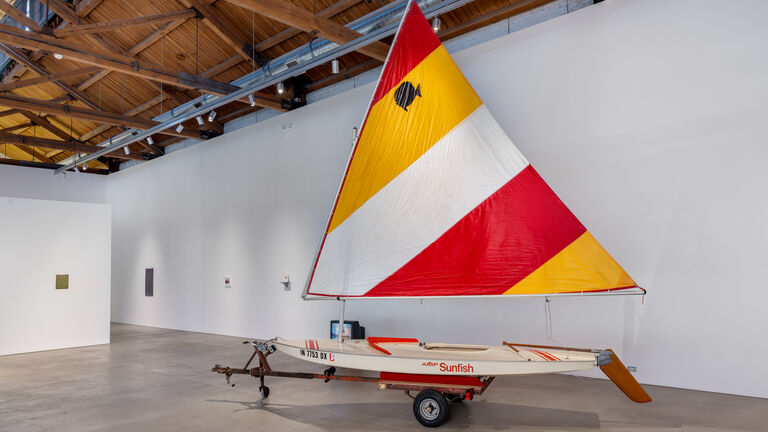A red, white, and yellow sailboat in a gallery space. The side of the boat has red text that reads "Sunfish."