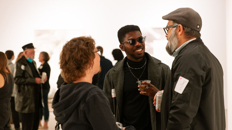 Three adults with deep and fair skintones converse in a gallery. 