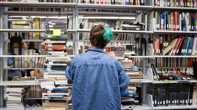 A student stands in front of a bookshelf in a library. 