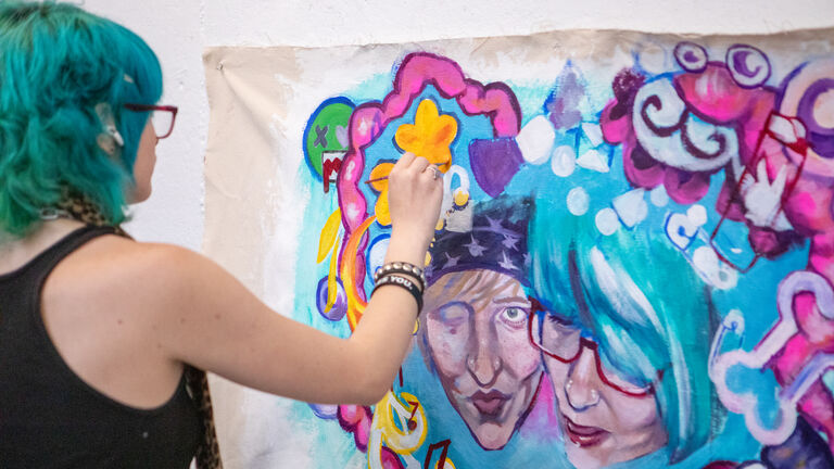 A person with a fair skintone and blue hair paints a colorful artwork of two faces in a heart of symbols. 