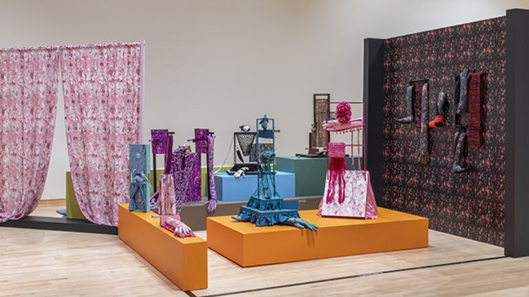 A gallery view of pieces of fiber art—bright, colorful curtains and a series of sculptures with hands in gloves peeking out from their bottoms