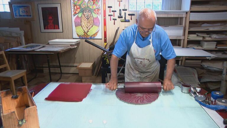 a bald man wearing an apron and blue shirt stands at their workstation, working on a print