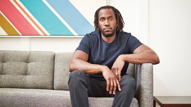 The artist Rashid Johnson poses on a couch in front of a brightly painted canvas