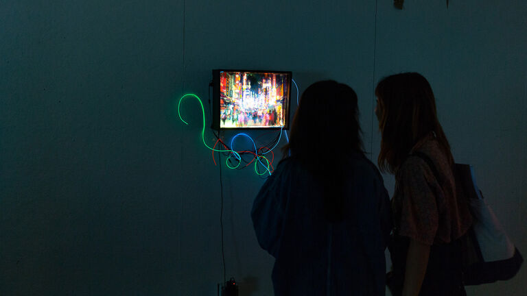 Two individuals in a dark room watch a screen with a cityscape with neon wires wrapped around it