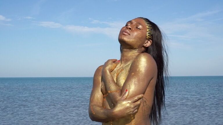 A Black woman stands nude in front of a large body of water, hugging herself with closed eyes. Gold clips line the hairline of her long, dark hair that reaches her lower back. Gold paint shimmers all over her body.