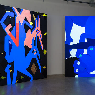 Two large paintings in a gallery. One depicts two creatures in blue and pink with bright yellow abstract shapes surrounding them and the other is a blue figure who is upside down. 