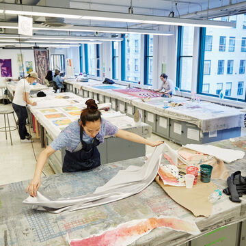Student in foreground spreading fabric on worktable. Several students working in studio on various textiles in the background.