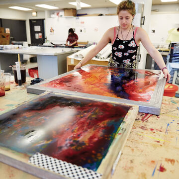 Student in foreground holding large screenprint covered in colorful ink. Other students in background working.