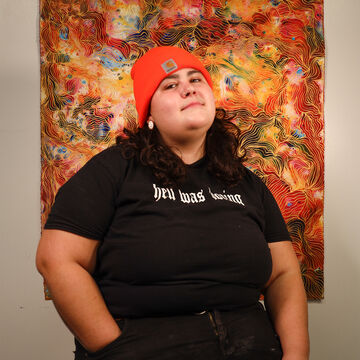 A person in an orange beanie and black outfit sitting in front of a colorful piece hanging on the wall