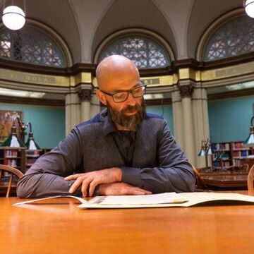 A bearded person reading a book in a large ornate library