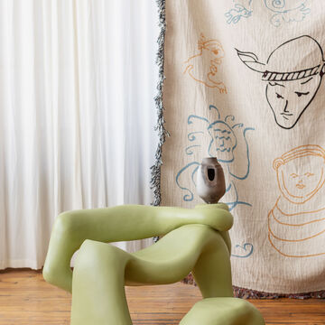 Three green abstract ceramic pieces resembling a chair and ottoman. There is a blanket with faces drawn on it behind it.