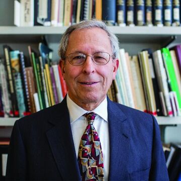 A bespectacled person in a suit smiling at the camera, with bookshelves behind them.