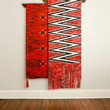 A large, hanging, red canvas featuring white and black zig zags with red fringe at the bottom. This piece is hanging over another red piece that is made out of reclaimed wood that features black plus signs with white tips.