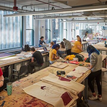 Students working individually in a sprawling studio space with large windows showcasing downtown Chicago architecture.