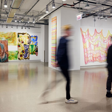 Patrons walking through galleries with bright pink and yellow textile hanging on the wall in foreground and painted textiles and canvases in background wall.