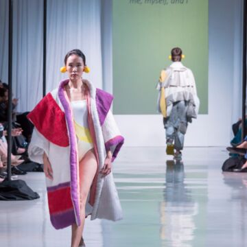 Runway show with model in foreground wearing a red, white, and purple cloak.