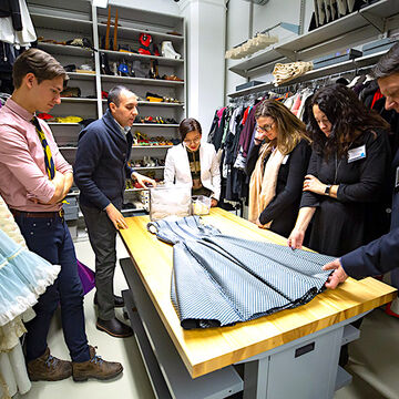 Several people looking at fabric laid out on a large table surrounded by garments in a fashion storage room