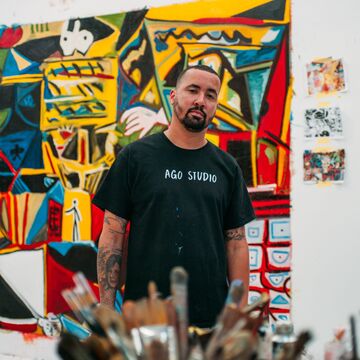 An artist stands in front of a colorful work of art in his studio