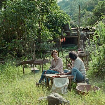 Two people sitting at a wooden table surrounded by trees and tall grass. 