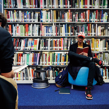A person reading in a bright blue chair with a large bookshelf behind them.