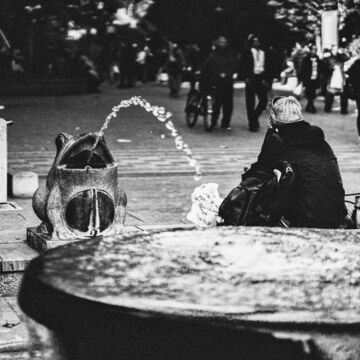 A black and white photograph of a person sitting by a fountain that is shaped like a frog. 