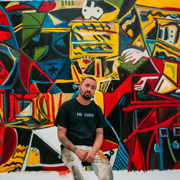 An image of Angel Otero sitting on a stool in front of a large abstract mural in his studio. 