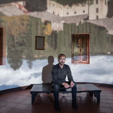 An image of Wafaa Bilal sitting in a large room with a scenic backgound projected on the walls.