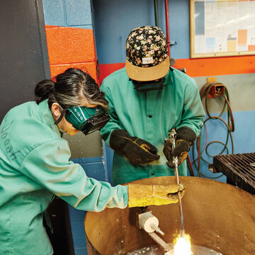 Two people wearing green protective smocks while heating up metal in a furnace.