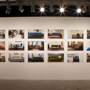 A large white wall in a gallery space with various pictures hung up. 