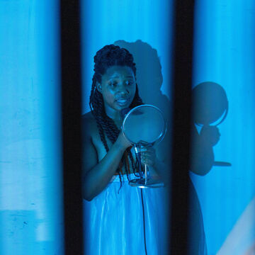 A person in a blue-lit room looks at a mirror while holding a microphone