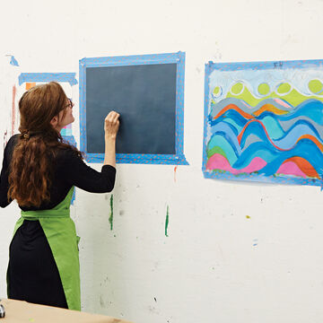 A teacher drawing something on a black piece of paper that has been taped to the wall.