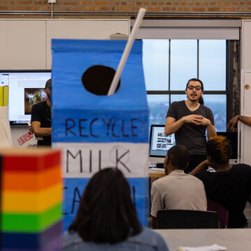 A student stands in front of a room next to a large blue and white art piece shaped like a milk carton while other students listen.
